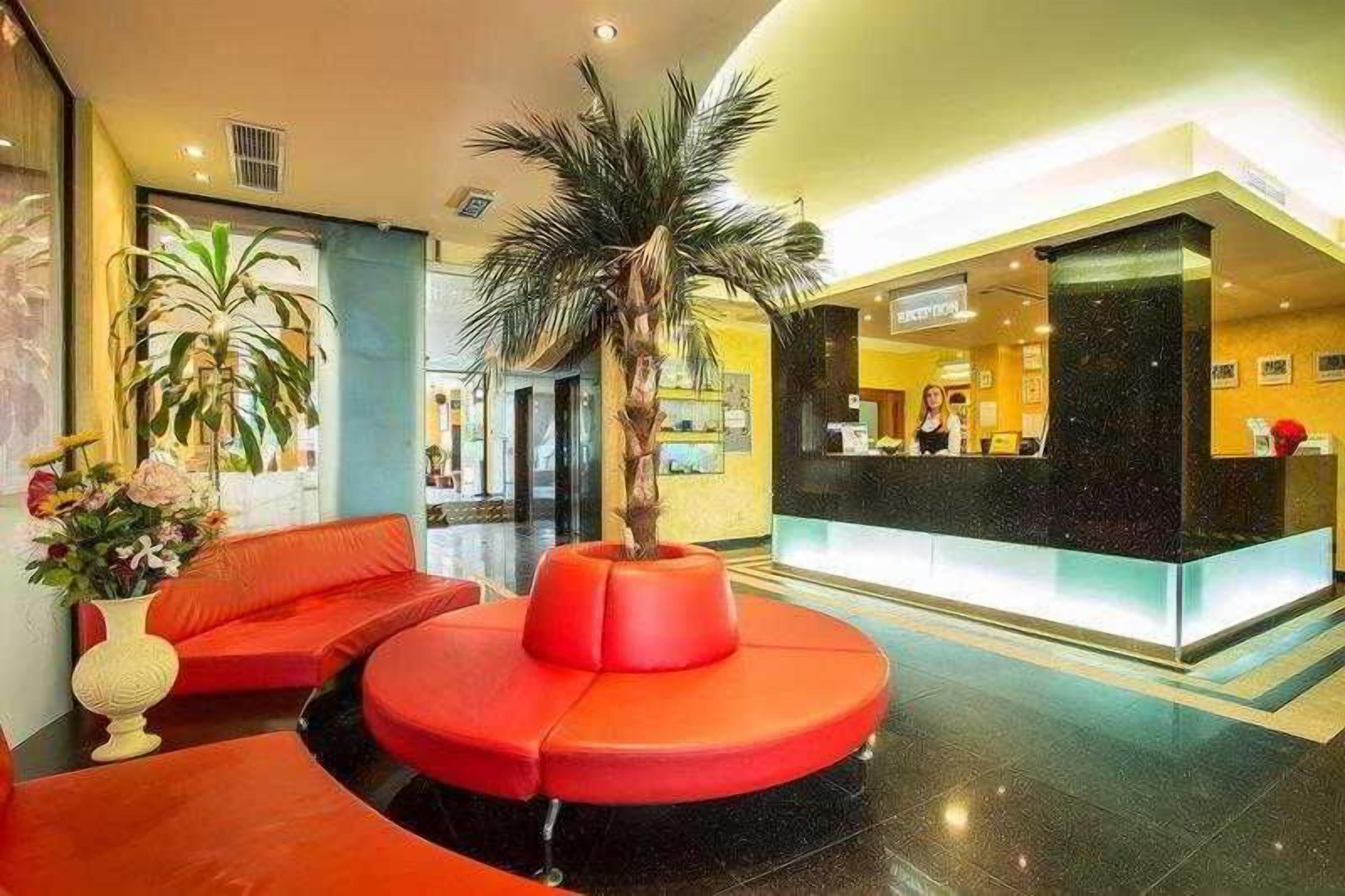 Un hall de hotel con mostrador de recepción, una zona de estar roja y una palmera.