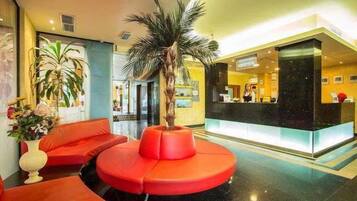 Un hall de hotel con mostrador de recepción, una zona de estar roja y una palmera.