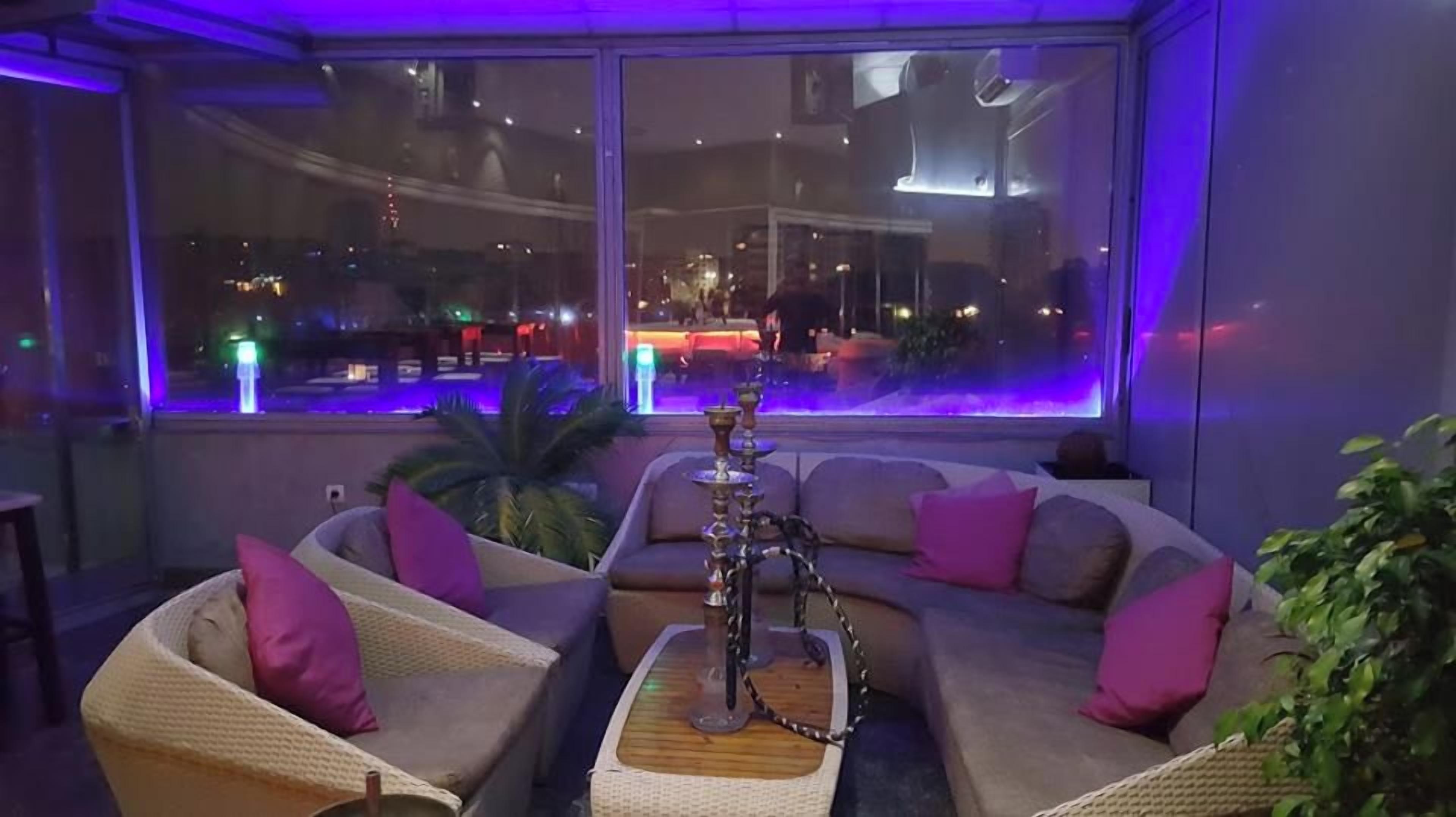A modern lounge area with a large window, cushioned seating, and a hookah.