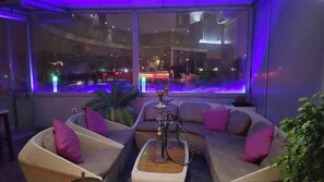 Un espacio lounge moderno con un amplio ventanal, asientos acolchados y un narguilé.