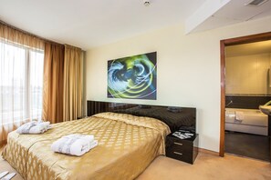 A hotel room with a bed, a bedside table with a magazine, a large abstract painting on the wall, and a bathroom visible through an open door. - Hotel Europe (Sofia)