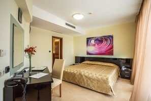 Una habitación de hotel con una cama, un escritorio con teléfono, una silla, un jarrón con flores, un cuadro en la pared y una puerta que da a otra habitación.