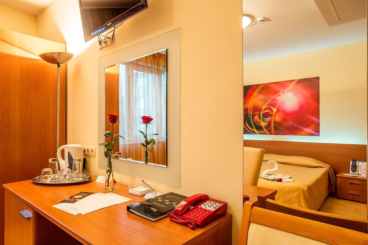 A hotel room with a wooden desk, a telephone, a mirror, a vase with a red rose, and a painting on the wall.