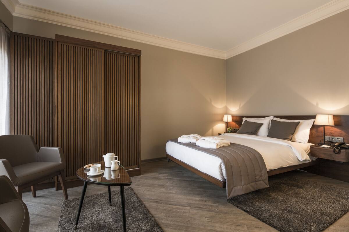 Ein modernes Hotelzimmer mit Bett, Nachttisch, Lampe und einer Sitzecke mit Tisch und Stühlen.