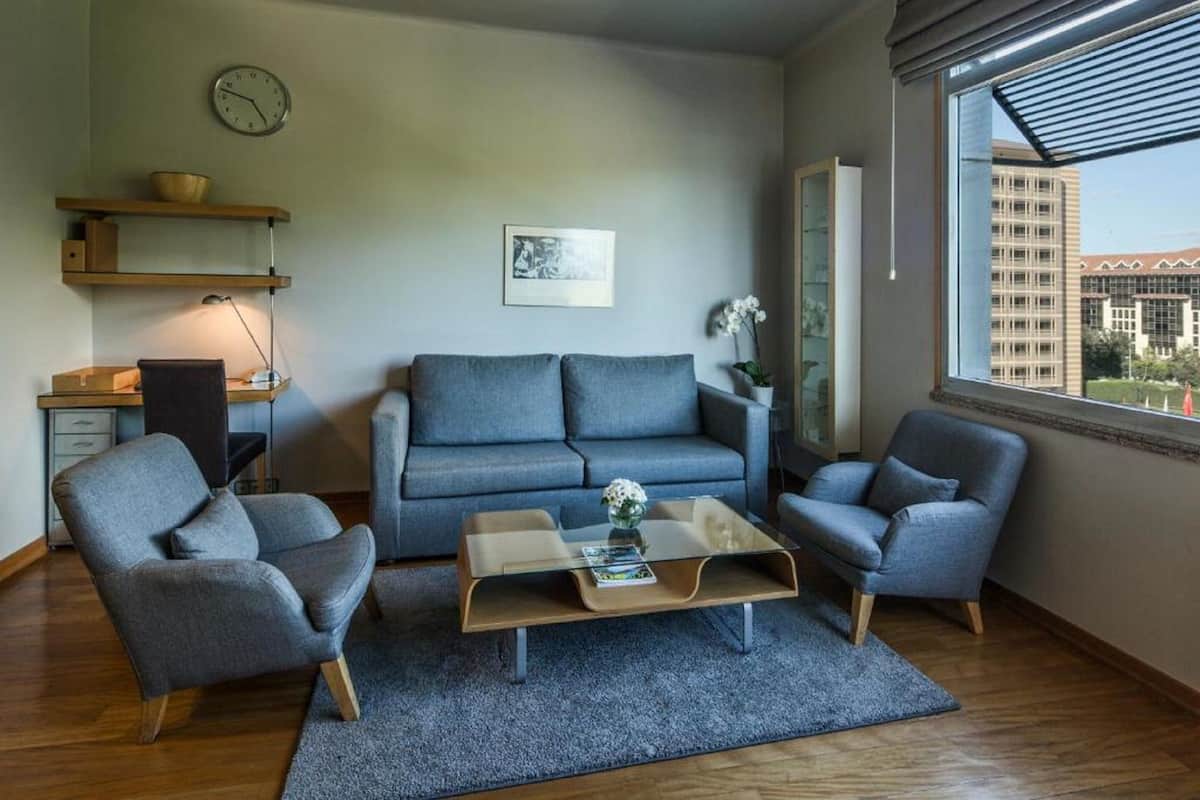 Ein modernes Wohnzimmer mit einem grauen Sofa, zwei Sesseln, einem Glastisch, einem Schreibtisch mit Stuhl, einer Uhr und einem Fenster mit Blick auf Gebäude.