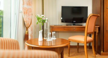 Una habitación de hotel con una mesa redonda de madera, un jarrón blanco con flores, un televisor, un escritorio de madera y una silla.