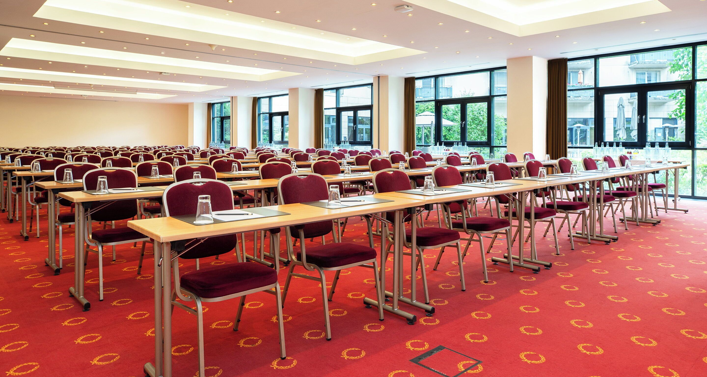 Un salón de conferencias con filas de mesas y sillas, ventanas amplias y una alfombra roja con diseño circular.