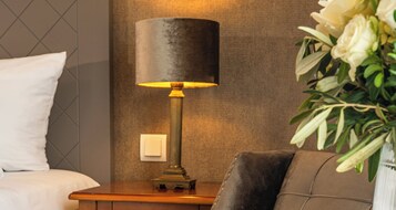Una camera d'albergo con un letto, un comodino con una lampada e un vaso con fiori.