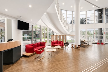 A spacious lobby with a reception desk, red seating, a staircase, and large windows.