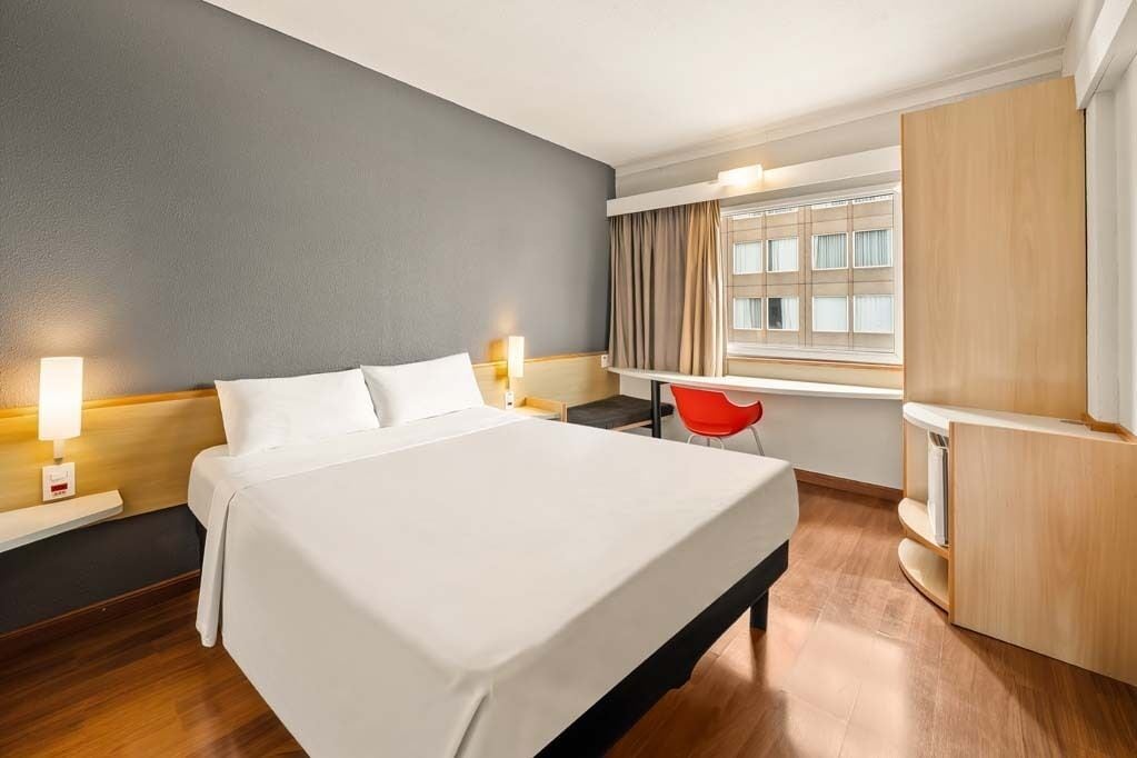 Quarto de hotel com cama, mesas de cabeceira, escrivaninha, cadeira, guarda-roupa e janela com cortinas.