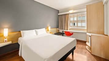 Quarto de hotel com cama, mesas de cabeceira, escrivaninha, cadeira, guarda-roupa e janela com cortinas.