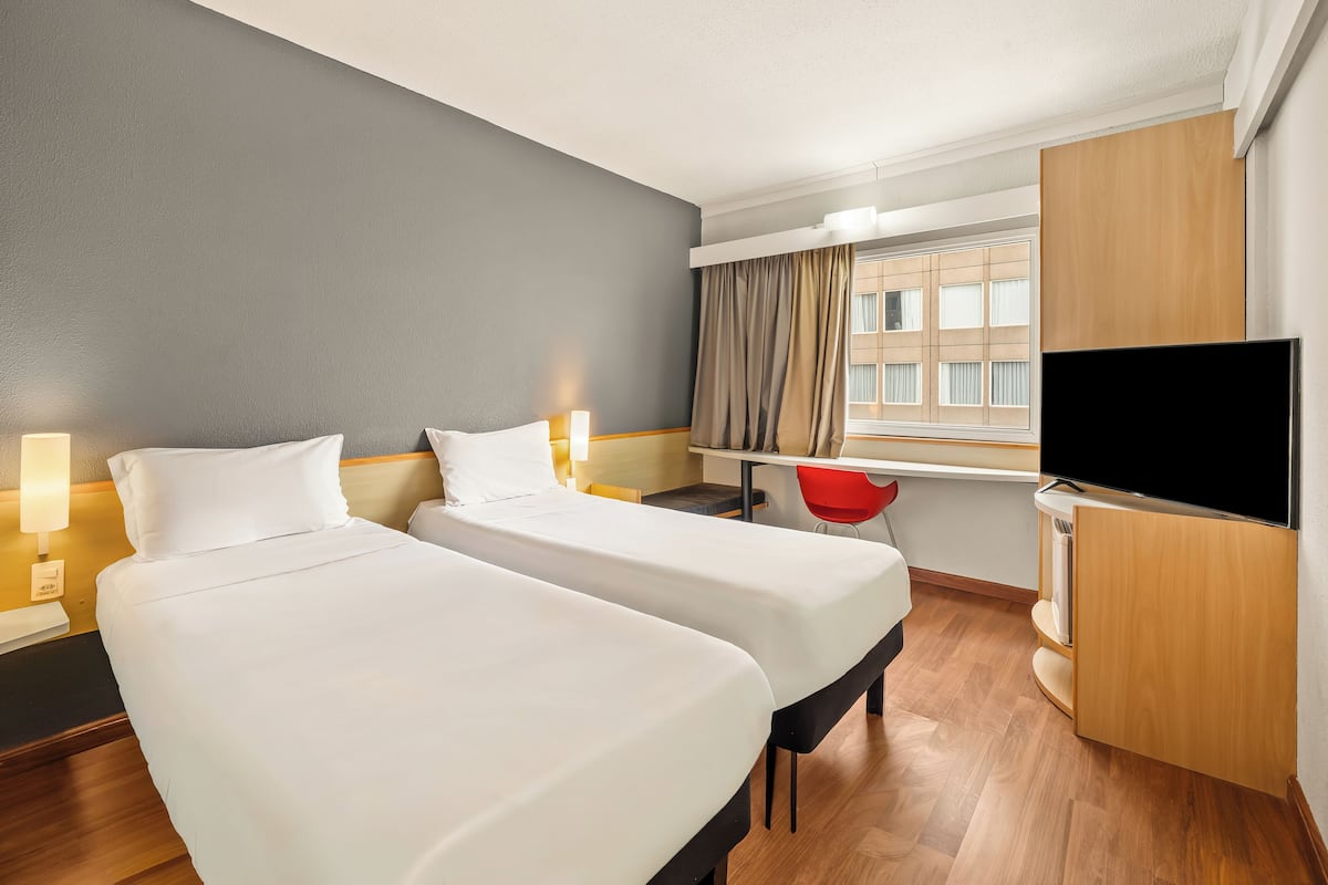 A hotel room with two beds, a desk, a chair, a TV, and a window with curtains.