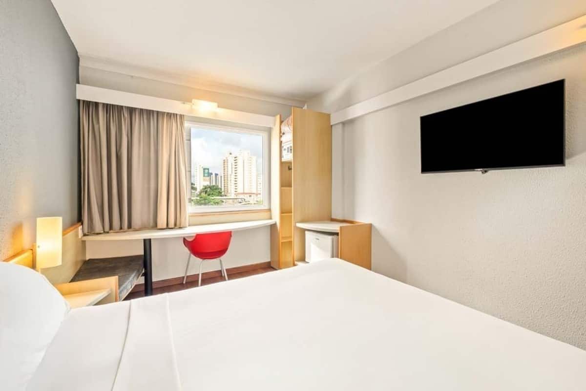 A hotel room with a bed, a desk with a chair, a TV mounted on the wall, and a window with curtains.