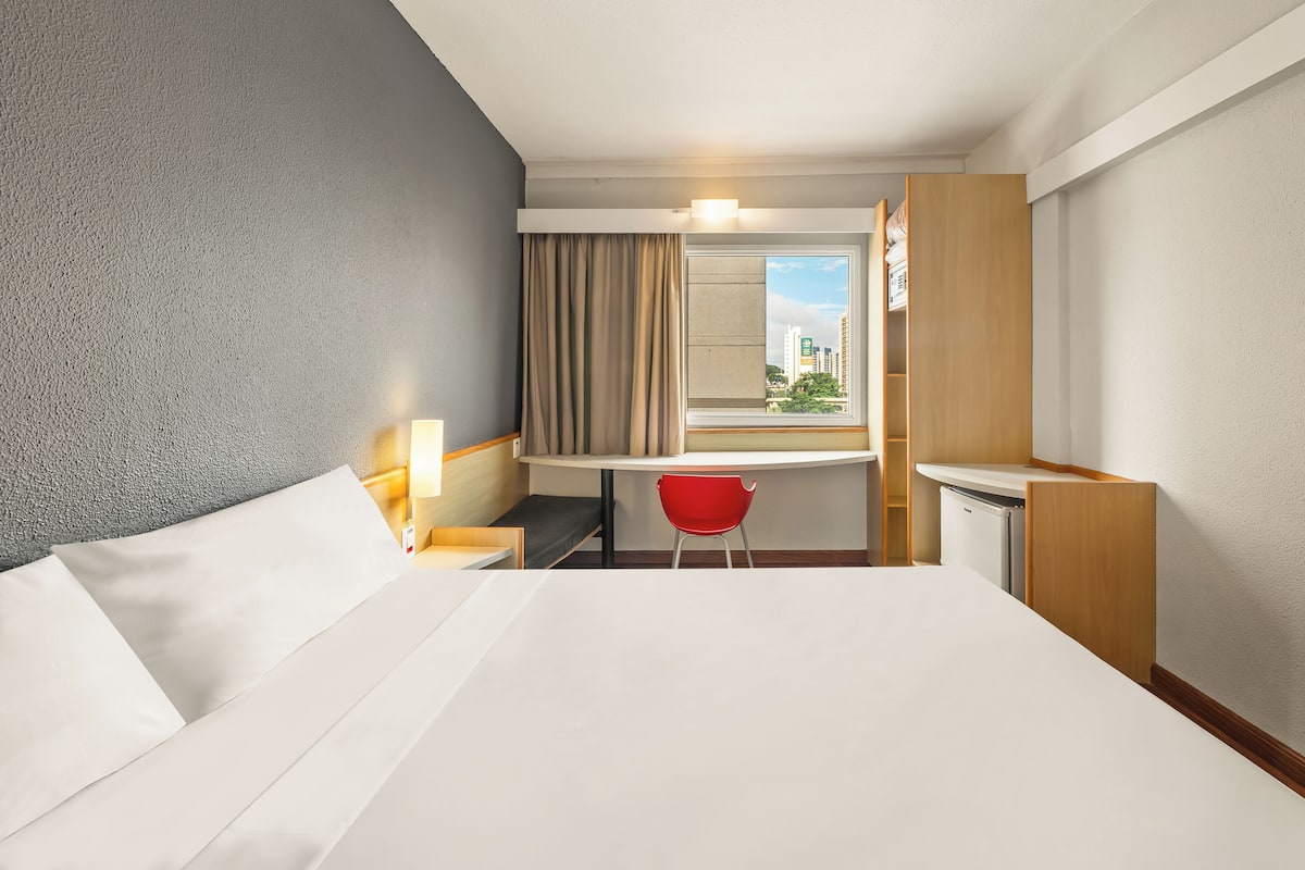 A hotel room with a bed, a desk, a chair, a window with curtains, and a view of buildings outside.