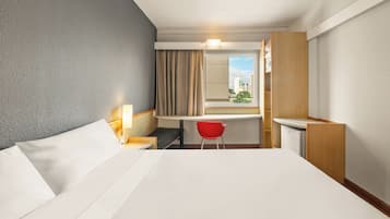Quarto de hotel com cama, escrivaninha, cadeira, janela com cortinas e vista para prédios.