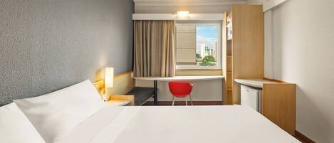 Habitación de hotel con cama, escritorio, silla, ventana con cortinas y vistas a edificios.