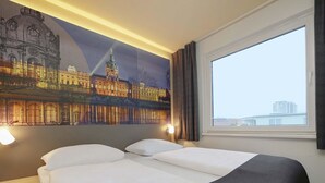 Une chambre d’hôtel avec un grand lit, une table de chevet, une lampe fixée au mur et une fenêtre donnant sur le paysage urbain.