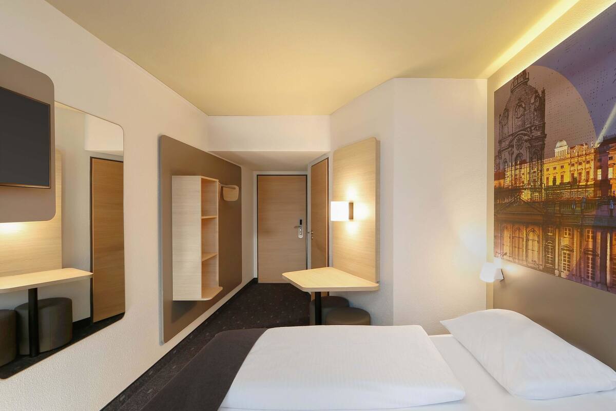 Habitació d'hotel amb llit, escriptori, mirall, armari i un quadre d'un paisatge urbà.