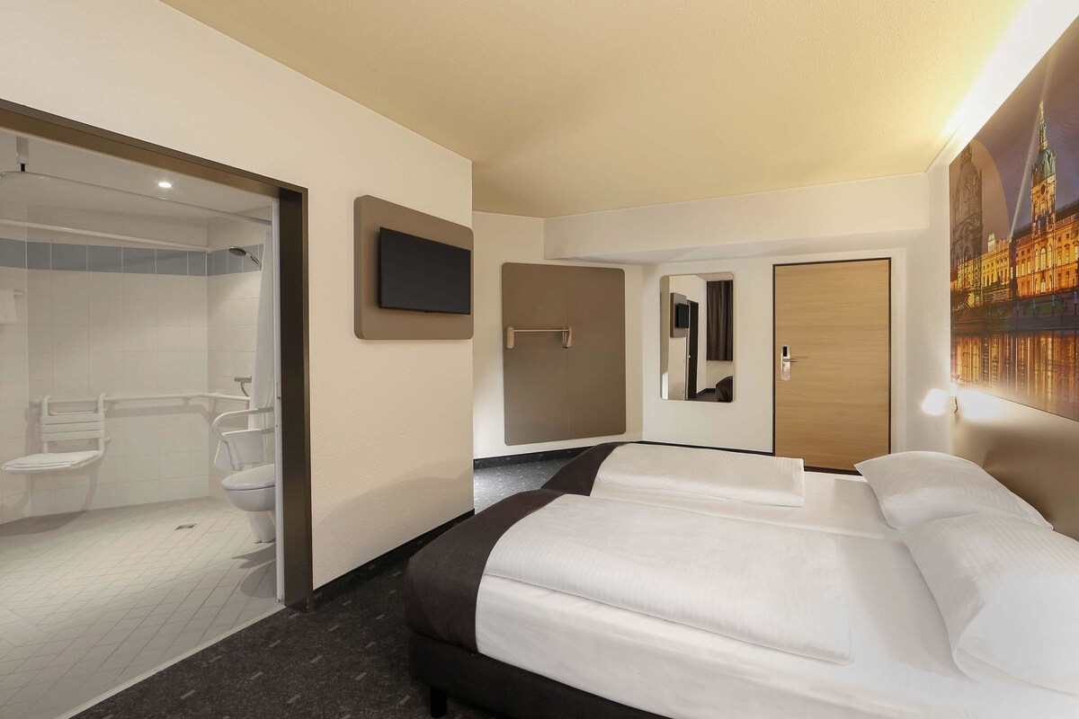 Une chambre d’hôtel comprenant un lit, un téléviseur fixé au mur, une salle de bain avec douche, toilettes et un miroir.