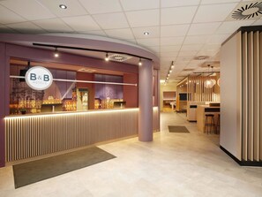 Un espace d’accueil au design moderne, comprenant un comptoir de réception, une grande fenêtre donnant sur le paysage urbain et une enseigne indiquant « B&B Hotels ».