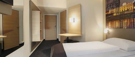 Habitación de hotel con cama, escritorio, silla, espejo, puerta y lámpara fijada a la pared.