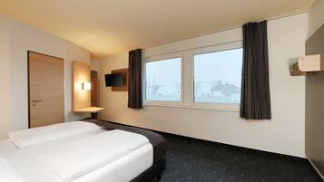 Ein Hotelzimmer mit einem Bett, einem an der Wand montierten Fernseher, einem Fenster mit Vorhängen und einer Tür.