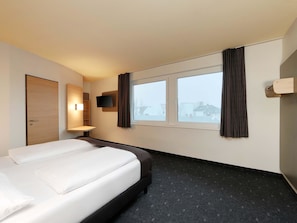 A hotel room with a bed, a TV mounted on the wall, a window with curtains, and a door. - B&B Hotel Berlin City-West (Berlin)