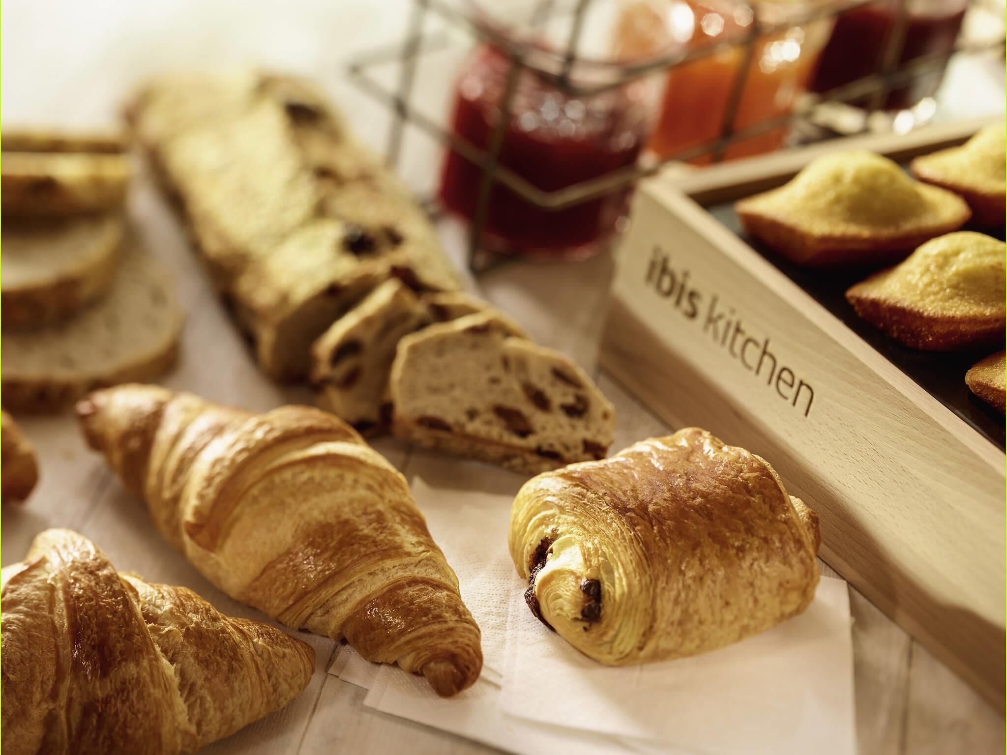 A variety of pastries and bread, including croissants and a baguette, displayed on a table with a label that reads 'ibis kitchen'.
