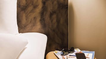 Une pièce avec un lit, une lampe fixée au mur, une paire de lunettes et un magazine sur une table.