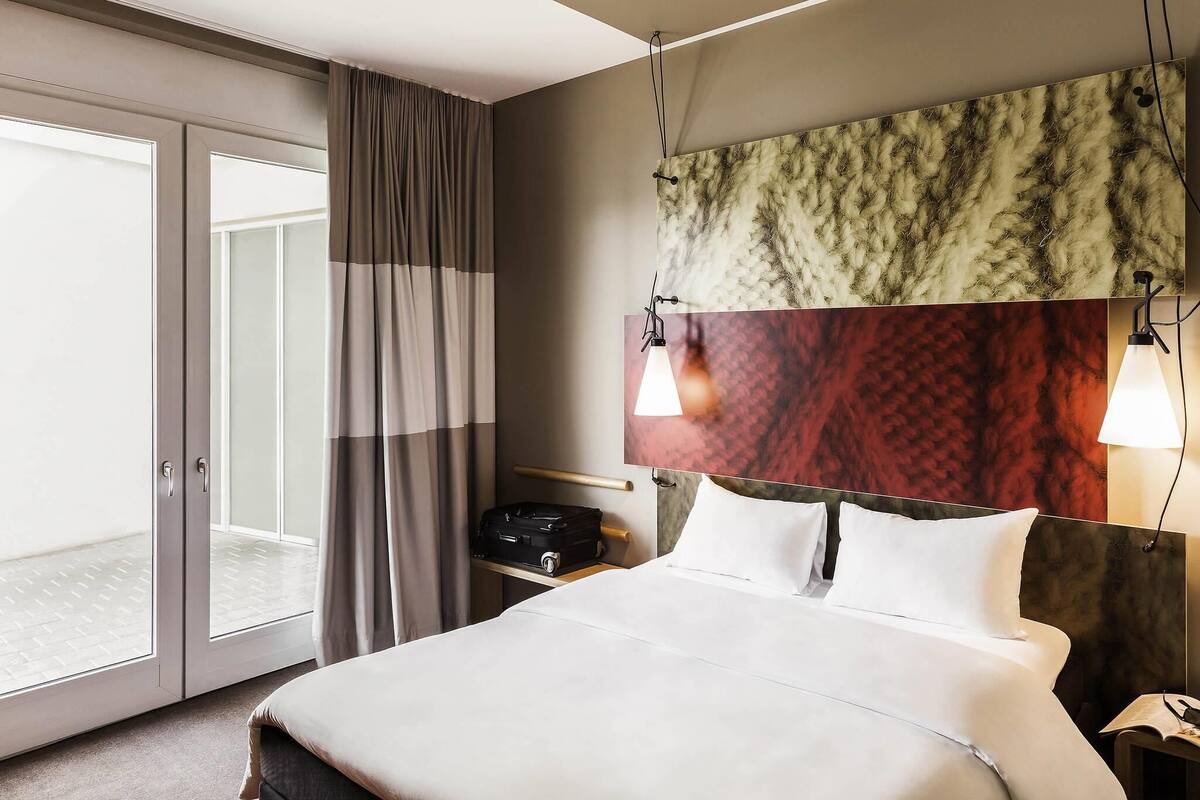 A hotel room with a bed, a suitcase, a nightstand, a lamp, and a view through a sliding door.