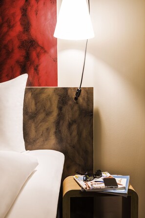 A neatly made bed with white linens, a bedside table with a lamp, and a magazine with sunglasses and a phone on it. - ibis Winterthur City (Winterthur)