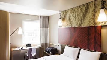 A hotel room with a bed, a desk with a laptop, a chair, a suitcase, and a window with curtains.