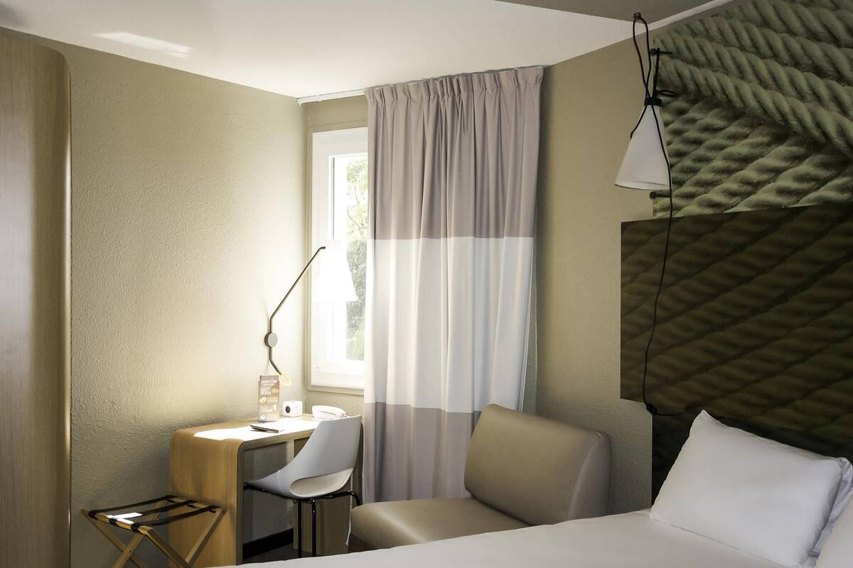 A hotel room with a bed, a desk with a chair, a lamp, and a window with curtains.