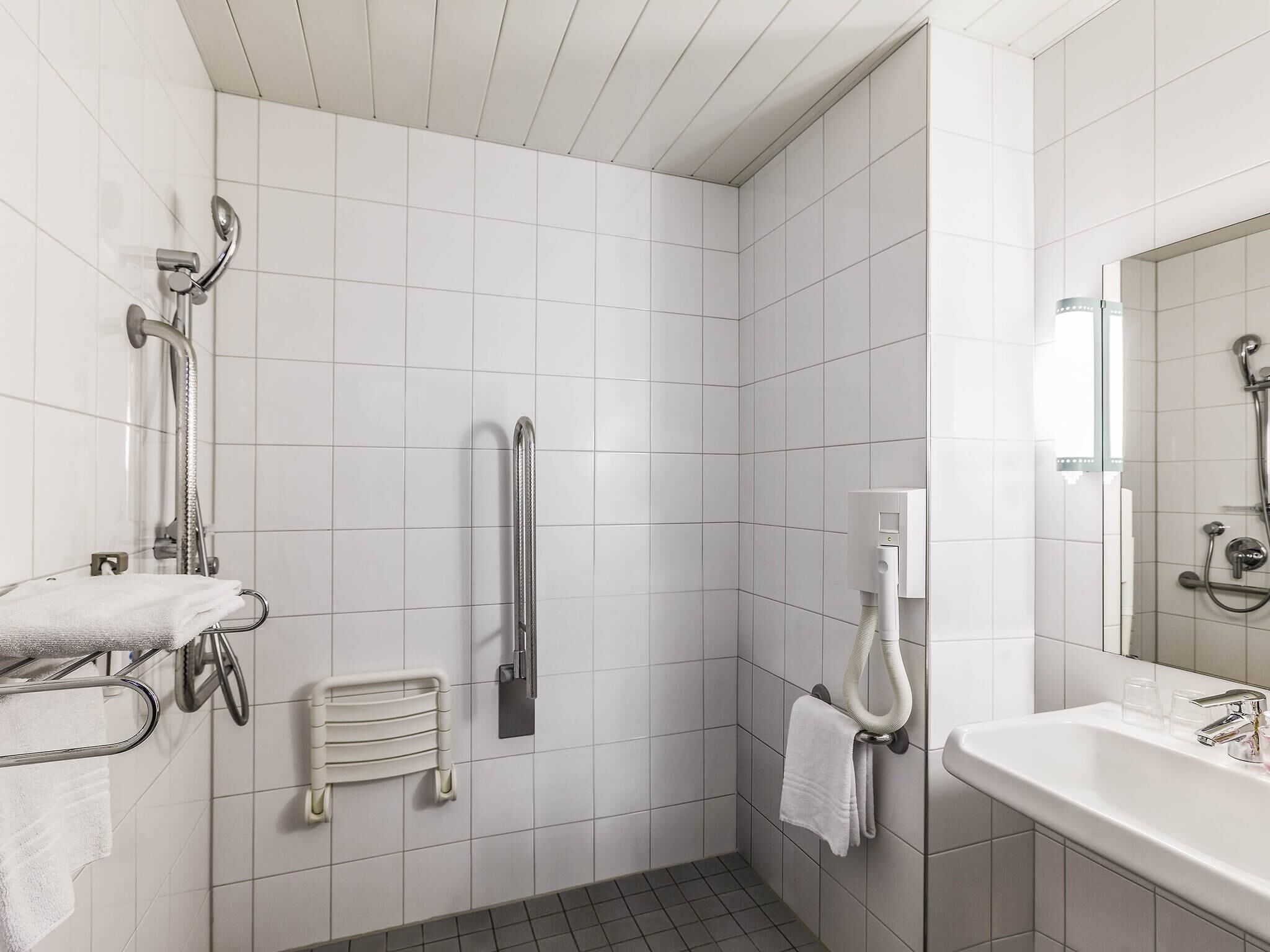 A modern bathroom with white tiles, a shower with a handheld showerhead, a grab bar, a towel rack, and a sink with a mirror.