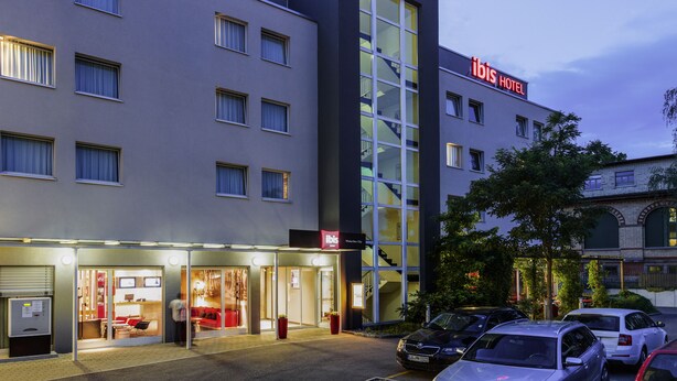 Un hotel moderno con una fachada de vidrio destacada, una escalera visible y un estacionamiento con varios automóviles.