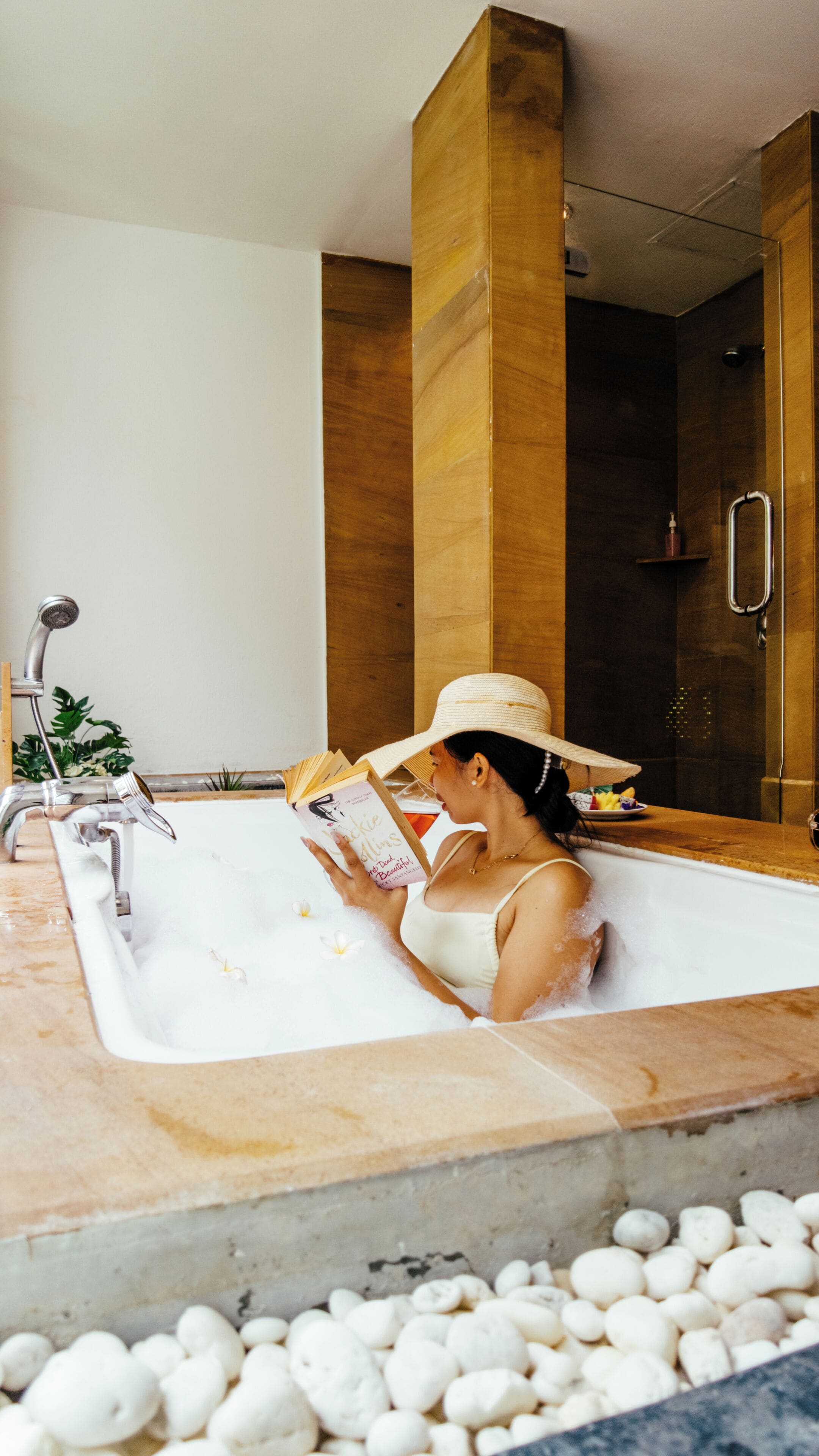 A woman in a bathtub reading a book, with a shower and a wooden column in the background.