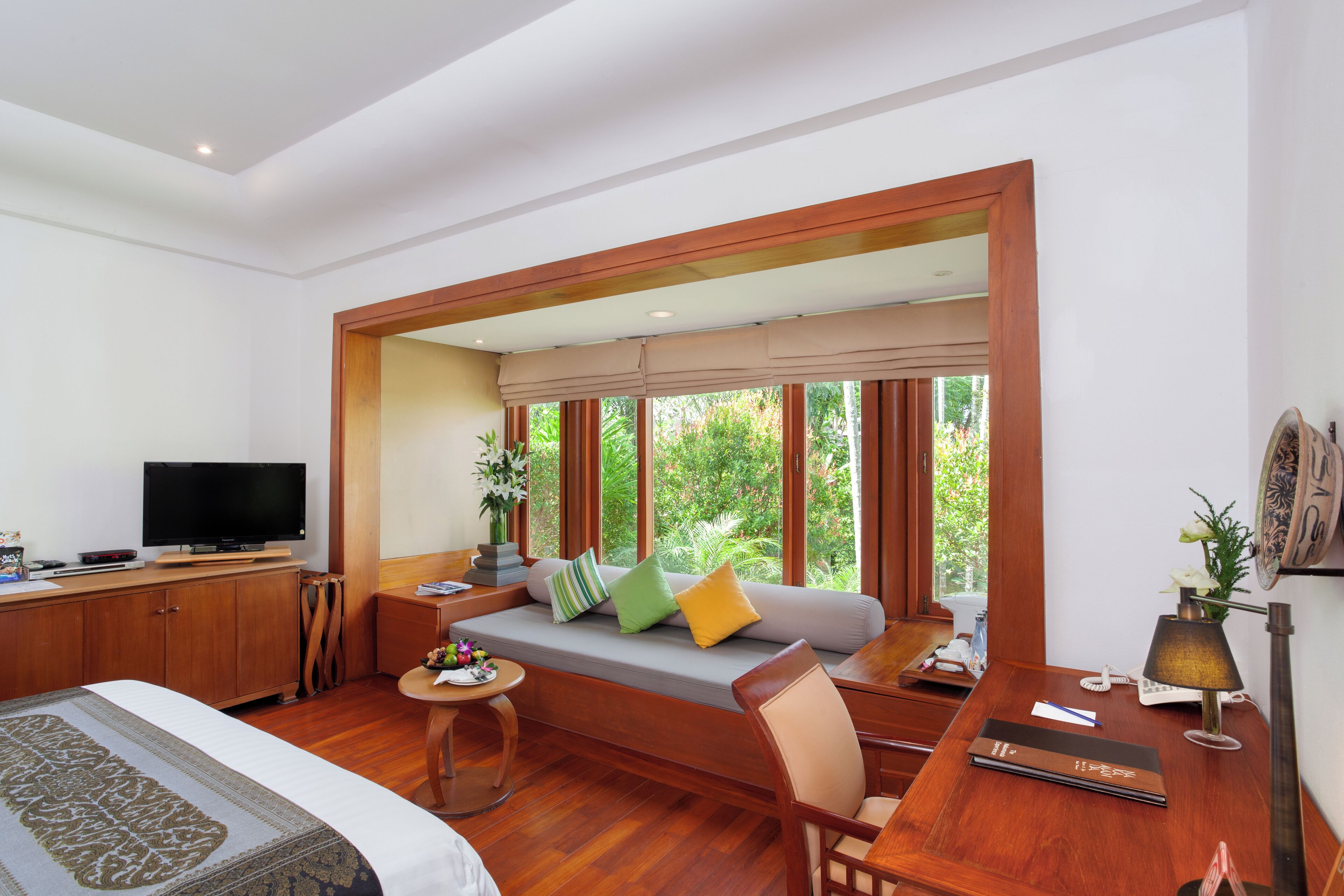 A room with a bed, a sofa with cushions, a television, a desk with a lamp, and large windows with a view of greenery.