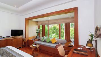 A room with a bed, a sofa with cushions, a television, a desk with a lamp, and large windows with a view of greenery.