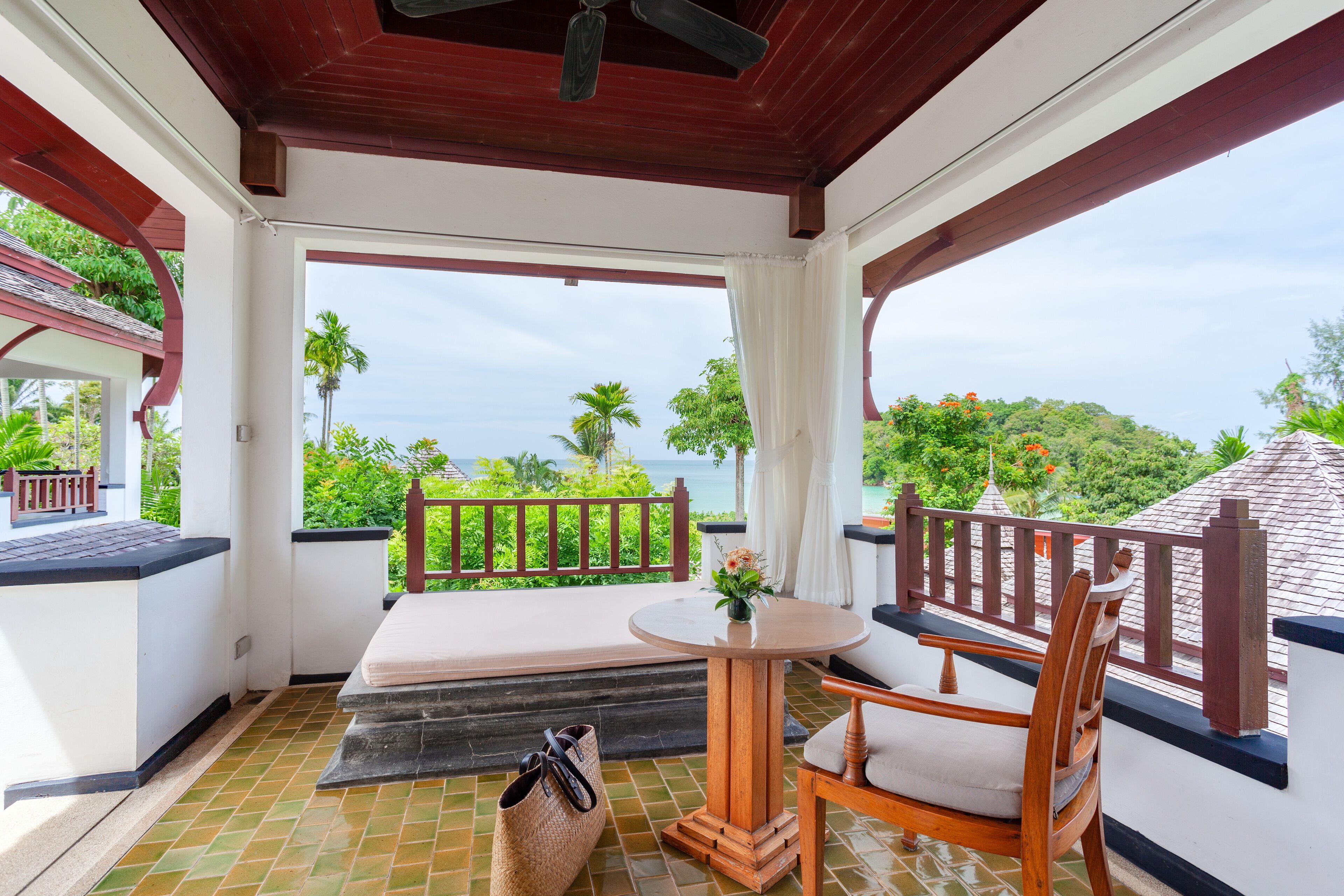A covered balcony with a wooden table and chairs, a cushioned bench, and a view of the ocean and greenery.