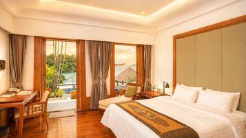 A hotel room with a large bed, a desk, a chair, and a view of a tropical landscape.