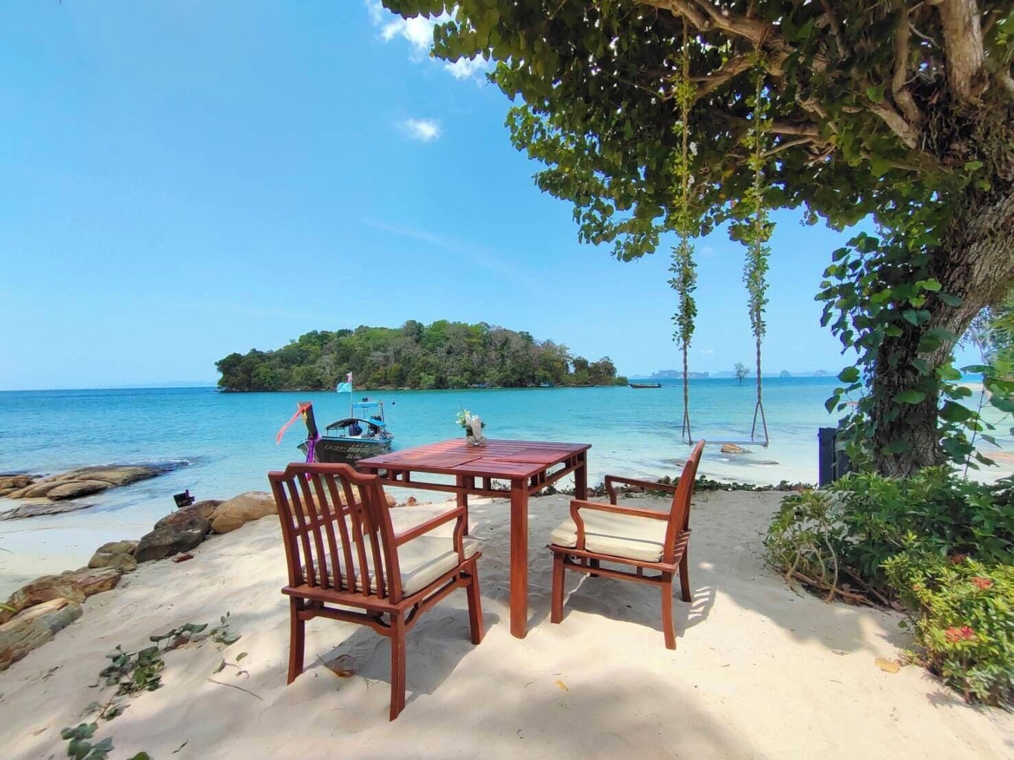 Una mesa y sillas de madera sobre una playa arenosa, con vista al mar y una pequeña isla al fondo.