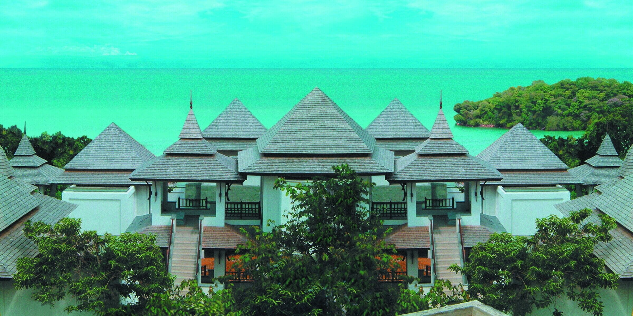 A hotel with multiple buildings, each featuring a steeply pitched roof and a balcony. The buildings are surrounded by lush greenery and a body of turquoise water in the background.