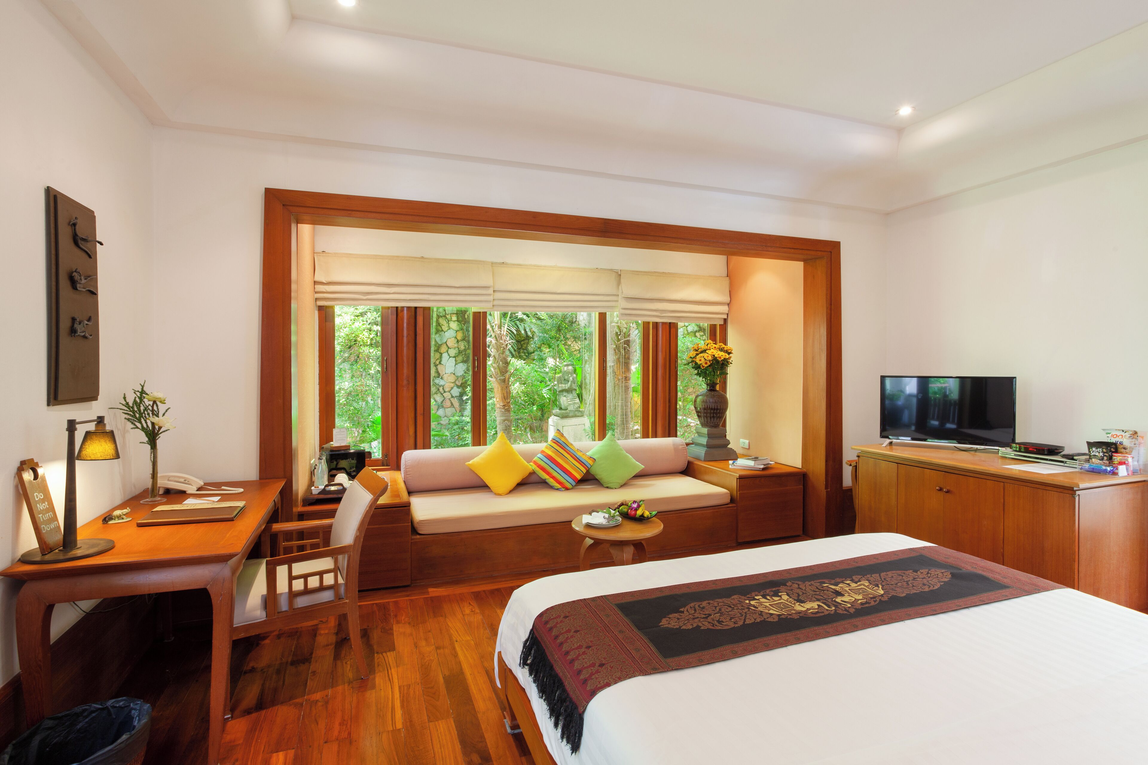 Une chambre d’hôtel comprenant un lit, un bureau, un canapé avec des coussins, une télévision et une fenêtre donnant sur un espace verdoyant.