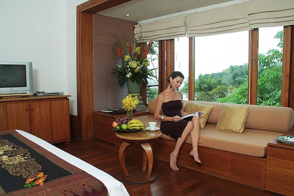 Una habitación con sofá, una pequeña mesa redonda con frutas y un florero, un televisor sobre un mueble y una ventana con vistas a la vegetación.