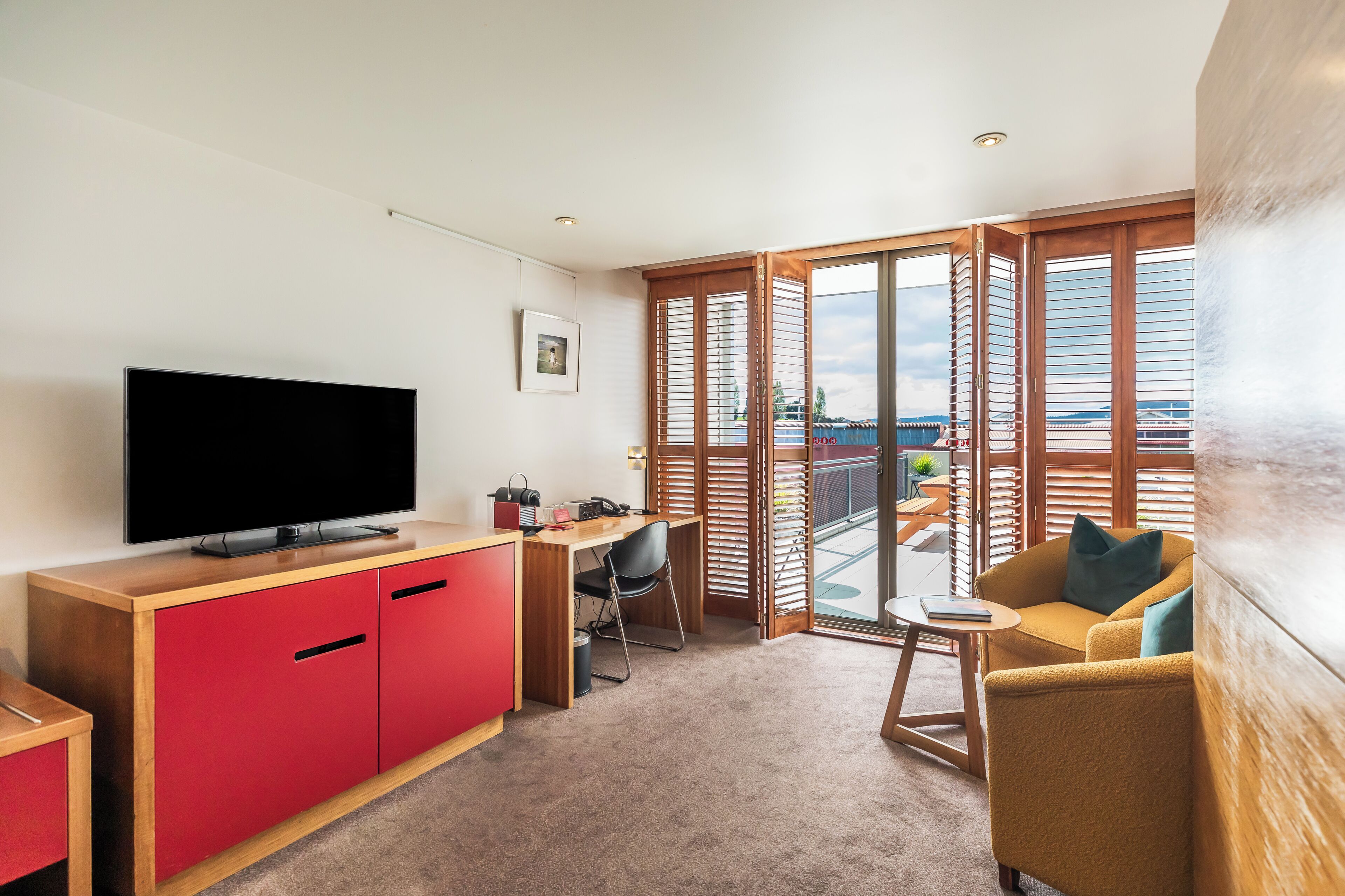 Una habitación de hotel moderna con televisión de pantalla plana, un escritorio de madera, un armario rojo, un sofá amarillo y un balcón con persianas de madera.