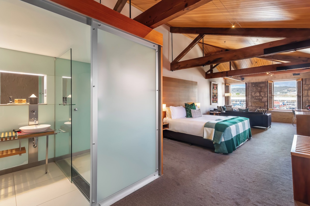 Habitación de hotel moderna con baño de vidrio, cama con colcha verde a cuadros y vistas de la ciudad a través de amplias ventanas.