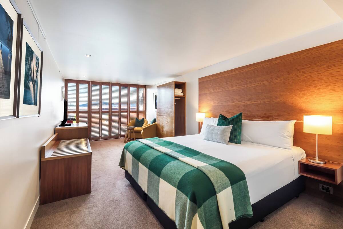 Une chambre d’hôtel moderne dotée d’un grand lit, d’une tête de lit en bois, d’un bureau avec une chaise, d’une fenêtre avec des stores et offrant une vue sur l’extérieur.