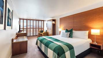 A modern hotel room with a large bed, a wooden headboard, a desk with a chair, a window with blinds, and a view of the outdoors.