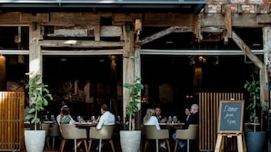 Een terras met buitenmeubilair, houten balken, plantenpotten en een schoolbord waarop staat dat het diner om 18.00 uur begint.