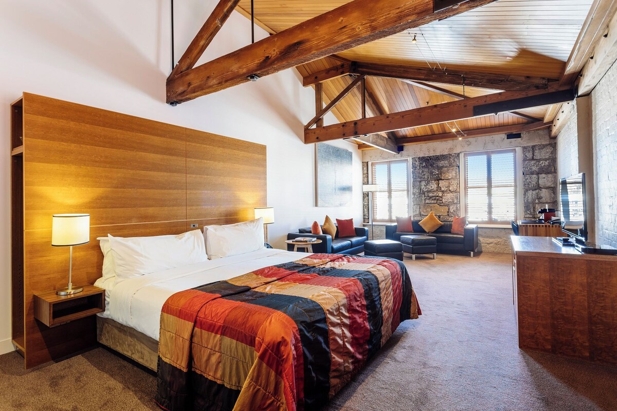 Une chambre d’hôtel avec une tête de lit en bois, un grand lit recouvert d’une courtepointe colorée, un bureau avec une chaise, un canapé et un mur en pierre.